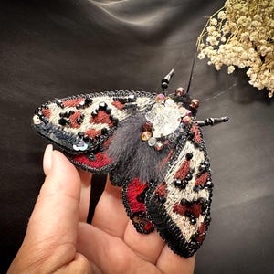 Puede incluir: Broche artesanal con forma de polilla. El broche presenta un patrón negro, rojo y beige en las alas, con abalorios y lentejuelas negras. El centro de la polilla tiene una textura de pelo gris oscuro y está adornado con cristales.