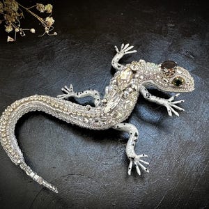Puede incluir: Una detallada figura de lagarto plateado, adornada con cristales y gemas brillantes. El lagarto tiene una cola larga y curvada y delicadas patas con manchas negras. Los ojos son oscuros, y el diseño es intrincado y elegante.