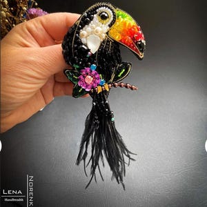 Puede incluir: Broche de tucán con cuentas con pico arcoíris, cuerpo negro y detalles blancos. El broche presenta una flor rosa y azul, plumas negras y está adornado con cuentas y perlas. El broche está hecho a mano.
