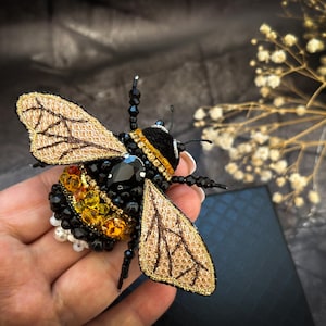 Puede incluir: Broche de abeja artesanal con intrincado trabajo de abalorios y alas bordadas. El broche presenta un cuerpo negro con detalles amarillos y naranjas, una gran gema negra y alas doradas. Las alas tienen un patrón texturizado.