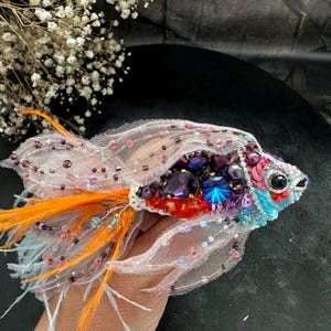 Puede incluir: Broche de pez hecho a mano con intrincados abalorios, lentejuelas y plumas. El broche presenta una combinación de colores púrpura, azul, naranja y rosa. El pez tiene un ojo detallado y está hecho con un material transparente similar a una red.