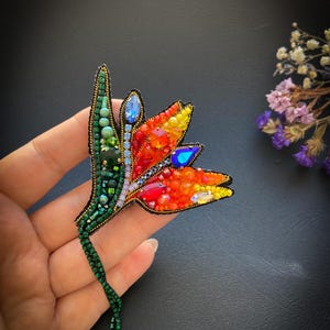Puede incluir: Broche de cuentas con forma de flor de ave del paraíso. El broche presenta colores vibrantes, incluyendo rojo, naranja, amarillo y azul, con hojas y tallo verdes. Está adornado con cuentas y cristales de varios tamaños.