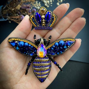 Puede incluir: Un broche de abeja artesanal y una pequeña corona, ambos adornados con cuentas y cristales azules y dorados. La abeja presenta un gran cristal iridiscente en su centro. La corona es una versión en miniatura del diseño de la abeja. El broche y la corona se muestran en una mano.