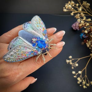 Puede incluir: Broche de polilla artesanal y ornamentado. El broche presenta un cuerpo de terciopelo y alas con intrincados abalorios en tonos azules, turquesas, rosas y dorados. Una gran gema azul adorna el centro. Un accesorio único.