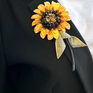 Puede incluir: Broche de girasol con pétalos amarillos y un centro de cuentas negras y doradas. El broche está sujeto a una blazer negra con una cinta y un tallo dorados.