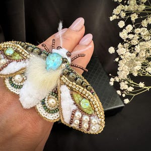 Puede incluir: Broche de polilla artesanal con intrincado trabajo de abalorios en oro, verde y blanco. Cuenta con una piedra preciosa turquesa central y está adornada con perlas. Las alas están detalladas con piel blanca.