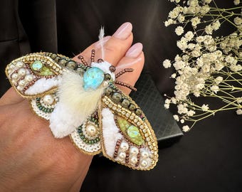 May include: A handcrafted moth brooch with intricate beadwork, featuring white, gold, and green accents. The brooch has a central turquoise gemstone and is embellished with pearls. The wings are detailed with white fur.