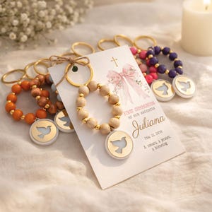 May include: A collection of keychains with wooden bead bracelets and dove pendants. A card reads "First Communion of our Daughter Juliana May 12, 2030". The keychains have gold-colored rings and are arranged on a white surface.