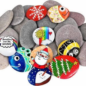 Puede incluir: Una colección de piedras de río lisas y ovaladas, algunas de color gris liso y otras pintadas a mano con diseños coloridos que incluyen un copo de nieve, un pez, un arcoíris con el texto "Somewhere over the RAINBOW", un sol, una abeja, Santa Claus y un árbol.