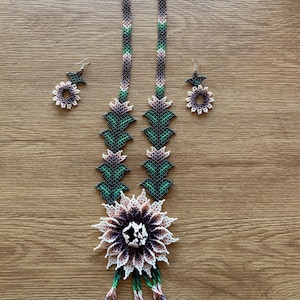 May include: A beaded necklace and matching earrings set. The necklace features a large, multi-colored flower pendant with a beaded design in shades of white, purple, and brown. The necklace and earrings have a floral design with green, purple, and white beads.