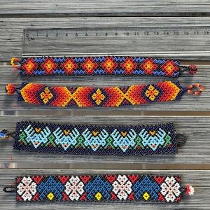 Può includere: Quattro braccialetti di perline intrecciate con motivi geometrici. Il braccialetto in alto presenta rombi rossi, arancioni e blu. Il secondo ha un design a chevron rosso, arancione e blu. Il terzo mostra motivi di uccelli blu su nero. Il quarto ha forme floreali blu e bianche.