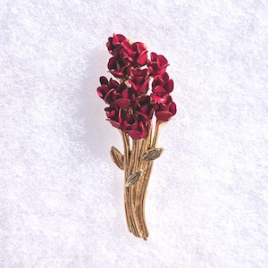 May include: A gold-toned brooch featuring a bouquet of deep red flowers. The floral arrangement is detailed with green leaves and is set against a white, textured background. The brooch is a vintage-style accessory.
