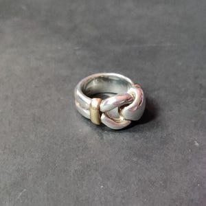 May include: Silver-toned ring with a knot design, featuring a gold-colored accent. The ring has a polished finish and a thick band. The knot design is the focal point, creating a unique and stylish accessory. The ring is set against a dark background.