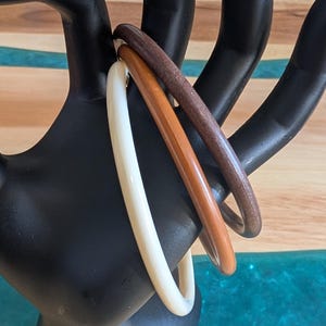 May include: Three bangles in ivory, light brown, and dark brown hues are displayed on a black hand-shaped jewelry holder. The bangles are smooth and appear to be made of wood or a similar material. The background is a wooden surface.