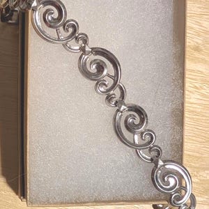 May include: Silver-toned bracelet with a repeating swirl design. The bracelet is displayed in a cardboard box with white padding. The swirls are connected by small, geometric links.