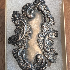 Pode incluir: Um broche antigo prateado com um design ornamentado e simétrico. O broche apresenta um espaço central retangular rodeado por detalhes de volutas, detalhes florais e um acabamento texturizado. É apresentado numa pequena caixa bege com acolchoamento branco.