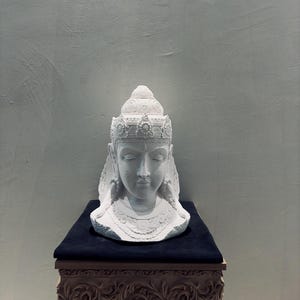 Puede incluir: Busto blanco de una figura serena con los ojos cerrados, adornado con un tocado y collar ornamentados. La escultura se exhibe sobre una superficie azul oscuro, encima de un soporte de madera tallada, sobre un fondo gris neutro.