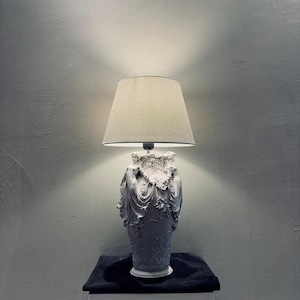 May include: A white decorative lamp with a beige lampshade. The lamp base is a detailed sculpture of figures and floral elements. The lamp sits on a black cloth atop a white column.