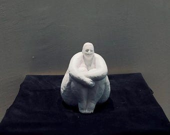 May include: A white sculpture of a person in a seated, curled position, with arms wrapped around the knees. The sculpture is on a black surface, atop a decorative wooden table. The background is a solid gray.