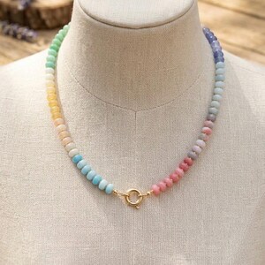 Peut inclure: Un collier de perles colorées présenté sur un mannequin. Le collier présente un dégradé de couleurs pastel, allant du vert au violet. Il est doté d'un fermoir doré.