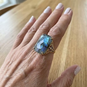 Könnte beinhalten: Ein Goldring mit einem großen, rechteckigen, facettierten Labradorit-Edelstein mit blauer und grauer Irisierung. Die Fassung ist mit filigranen, durchbrochenen Metallverzierungen an den Seiten der Ringschiene versehen.