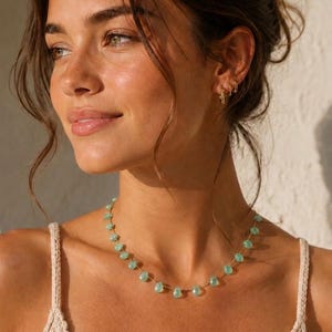 Può includere: Una collana delicata caratterizzata da una serie di piccole pietre preziose verde chiaro a forma di goccia, distanziate uniformemente lungo una sottile catena color oro. La collana poggia sul collo, esaltando la qualità delle pietre.
