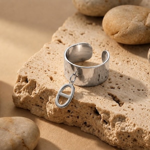 Könnte beinhalten: Ein offener Ring aus poliertem silberfarbenem Metall mit einem breiten, glatten, kreisförmigen Band und einem hängenden Anhänger in Form eines horizontalen Ovals mit einem vertikalen Mittelsteg, befestigt durch einen kleinen Ring.