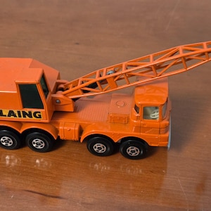 May include: Orange toy crane truck with the word "LAING" printed on the side. The crane has a long, articulated arm with a hook. The truck has six black wheels and a detailed cab.