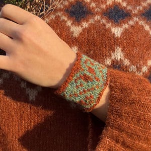 May include: A knitted cuff bracelet in shades of orange, green, and red. The cuff features a geometric pattern and is worn on a person's wrist. The sweater in the background has a diamond pattern.
