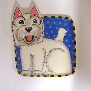 May include: A ceramic West Highland White Terrier-themed decorative tray. The tray features a white dog with a pink tongue, set against a blue background with white polka dots. The tray's border is black and yellow checkered.