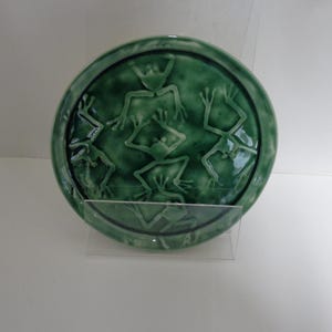 May include: A round, green ceramic plate with a raised relief design of stylized frogs. The plate has a glossy finish and is displayed on a clear stand. The frogs are arranged in a circular pattern.