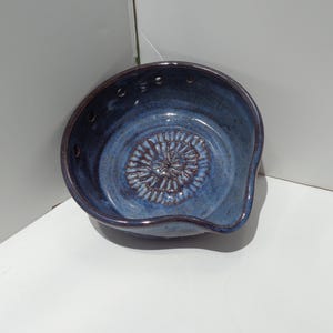 May include: A handmade, blue ceramic grater bowl with a dark rim and a textured interior. The bowl features a built-in grater in the center and small holes around the rim. The bowl is designed for kitchen use.