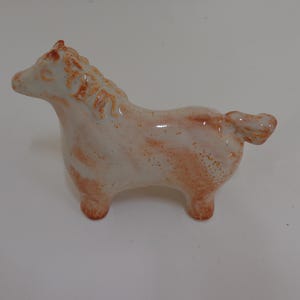May include: A ceramic horse figurine in a standing pose. The horse is primarily white with reddish-brown accents on the mane, body, and legs. The figurine has a smooth, glossy finish and is a decorative item.