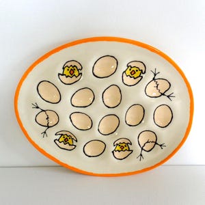 May include: Oval ceramic serving platter with an orange rim. The white interior features egg-shaped indentations, some with painted yellow chicks hatching from cracked eggs. The eggs are a light beige color, and the outlines are black.