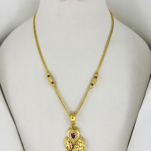 May include: A gold-colored jewelry set featuring a necklace and matching earrings. The necklace has a pendant with a heart-shaped red stone and a textured design. The earrings have a similar design with a red stone and a textured gold finish.