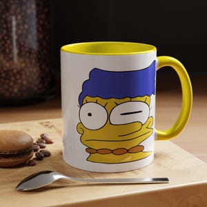 May include: White ceramic mug with a yellow interior and handle, featuring a cartoon image of Marge Simpson winking. The mug is on a wooden surface with a spoon and a pastry.