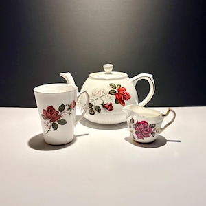 May include: A white porcelain tea set comprising a teapot, a creamer jug, and a teacup. Each item is adorned with a red rose floral design and gold edging. The teapot has a lid and a curved handle. The creamer has a small handle. The teacup has a handle.
