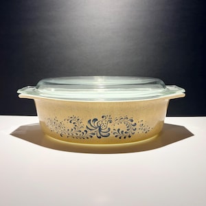 May include: A vintage casserole dish with a clear glass lid. The dish is a speckled tan colour with a blue floral design around the sides. The dish is designed for cooking and serving.