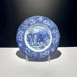 May include: A round ceramic plate featuring a blue and white transferware pattern. The centre depicts a winter scene with figures on horseback, while the wide rim is decorated with a detailed border of various flowers, leaves, and foliage.