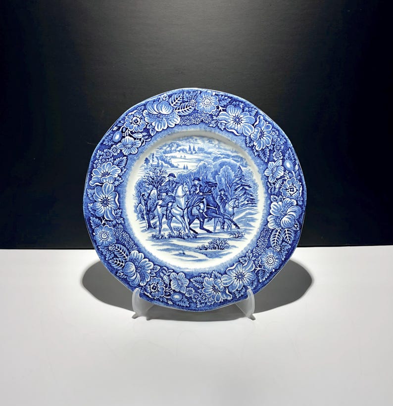 May include: A round ceramic plate featuring a blue and white transferware pattern. The centre depicts a winter scene with figures on horseback, while the wide rim is decorated with a detailed border of various flowers, leaves, and foliage.