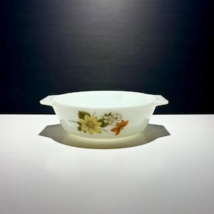 May include: A white, oval casserole dish with two handles. The dish has a floral design with yellow, white, and orange flowers. The dish is made of a translucent material and sits on a white surface.