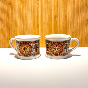 May include: Two off-white ceramic mugs featuring a repeating horizontal band of orange, brown, and yellow geometric patterns. Each mug has a curved handle on the side and a speckled texture throughout the base material of the ceramic body.