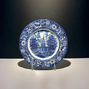 May include: A round ceramic plate featuring a blue and white transferware pattern. The centre depicts a historic building with a tall steeple, trees, and figures. The wide border is decorated with a dense floral motif of blossoms and leaves.