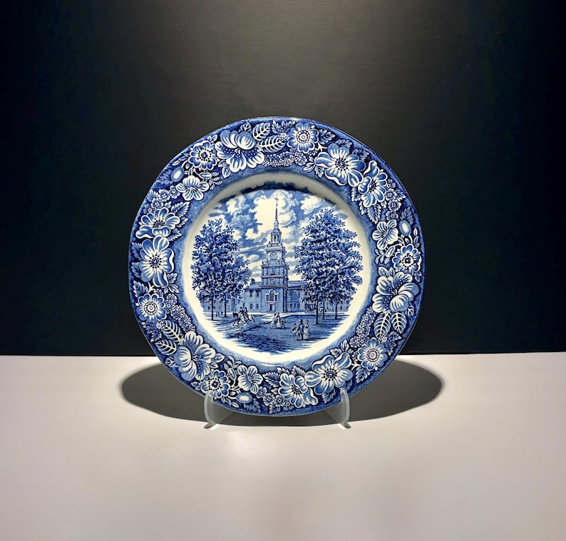 May include: A round ceramic plate featuring a blue and white transferware pattern. The centre depicts a historic building with a tall steeple, trees, and figures. The wide border is decorated with a dense floral motif of blossoms and leaves.