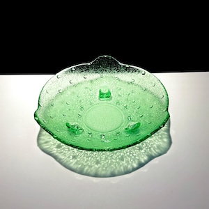 May include: A green glass decorative bowl with a textured surface and a bubble-like pattern. The bowl has a scalloped edge and three small feet. The bowl is approximately 20 cm in diameter.