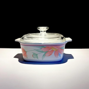 May include: A rectangular white casserole dish with integrated side handles and a clear glass lid. The exterior features a watercolour-style floral pattern with pink and orange petals, green leaves, and a thin pink horizontal stripe near the rim.
