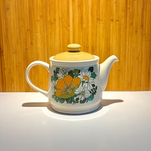 May include: A white ceramic teapot featuring a floral pattern with large orange and white flowers surrounded by green leaves. The teapot has a curved handle, a short spout, and a speckled tan lid with a small knob handle on the top centre.