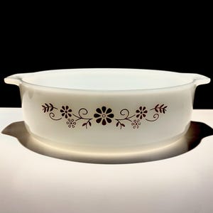 May include: A white round glass casserole dish with integrated side handles. The exterior features a brown floral pattern consisting of stylized flowers, leaves, and swirling vines centered on the side of the bowl against the opaque white glass.