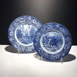 May include: Two round ceramic plates featuring blue and white transferware patterns. The left plate depicts a colonial building with a steeple, while the right plate shows figures on horseback in a snowy landscape. Both have floral borders.