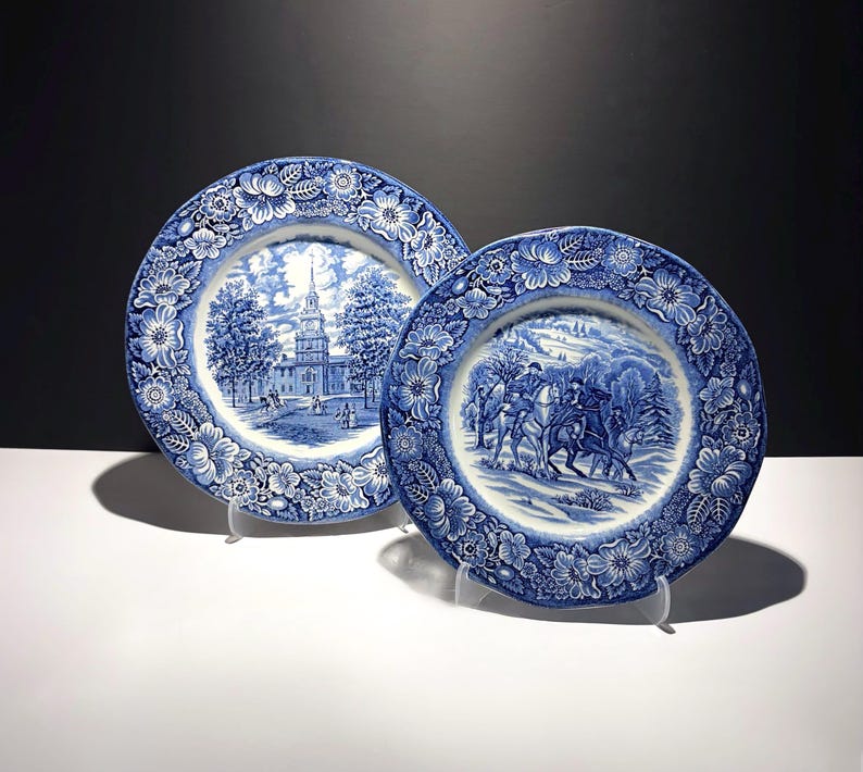 May include: Two round ceramic plates featuring blue and white transferware patterns. The left plate depicts a colonial building with a steeple, while the right plate shows figures on horseback in a snowy landscape. Both have floral borders.